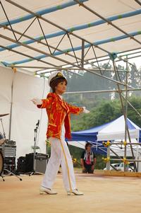 赤いジャケットと帽子を身につけた女性が、ステージ上で両手を広げてパフォーマンスしている写真