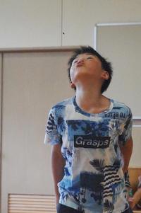 青い柄のシャツを着た男の子が、顔を上に上げて真剣な表情でスイカの種を飛ばしている瞬間の様子の写真