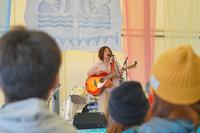 ギターを持ったアーティストが屋内ステージで歌っており、観客が手前から見守っている、ライブ演奏の写真