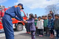 消防車の前で、向かい合った消防士と女の子が何かを手渡している様子を、周りの園児たちが見守っている写真