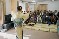 和服を着た女性が扇子を持って舞を披露し、それを座布団に座った大勢の観客が見守っている室内の写真