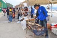 テント下で男性が炭火で魚介類を焼いており、来場者が並んで買い物している、屋外の屋台の写真