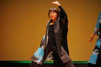 舞台の上で黒と青の和風衣装をまとった女性が、真剣な表情で片手を高く上げて力強く踊っている伝統舞踊の一場面の写真