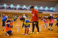 体育館の中で赤い服を着たコーチが、黄色とオレンジ色のビブスを着た子どもと体を使った準備運動をしている様子の写真