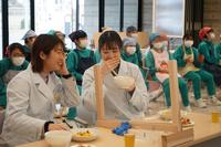 透明なアクリルボードが設置された席に白衣を着た2人の女子学生が座り、談笑しながら食事をしている写真