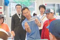 青いシャツを着た男性がトビウオを両手で持ち参加者達に見せている様子の写真