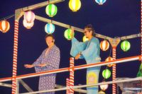 提灯が並ぶやぐらの上で、2人の浴衣姿の男性が盆踊りを踊っている写真