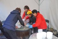 テントの中で女性たちが大きな鍋を囲み、白い器に料理を盛り付けている写真