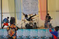 バンド演奏中に一人のメンバーがジャンプして宙に浮いている瞬間が撮影された、躍動感あふれるライブステージの写真