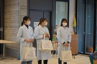 白衣を着た3人の女子大学生がイラストが描かれた白い手提げバッグをそれぞれ持ち、横一列に並んで立っている写真