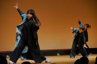 黒と青の和風衣装を身にまとった数人の女性たちが、舞台上で腕を大きく広げて力強く踊っている伝統舞踊の一場面の写真