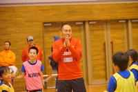 体育館の中で赤い服を着たコーチが笑顔で手を合わせて話しかけている様子を、周りの子どもたちが見ている場面の写真