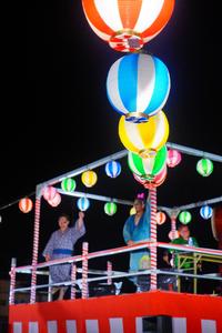 カラフルな提灯が輝くやぐらの上で、浴衣姿の人たちが左手を上げながら踊っている写真