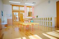 明るい日差しが差し込む保育園の教室で、中央に黄色いテーブルと椅子が整然と並び、右側には収納棚が設置され、奥には大きな窓とカーテンが見える写真