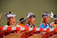 鉢巻きを締めた子どもたちが、色鮮やかな衣装を身に着けて真剣な表情で太鼓を力強く叩いている演奏風景の写真