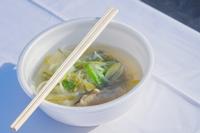 ネギやと貝が入った温かい汁物が、使い捨ての容器に盛られ箸が添えられている写真
