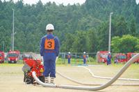 ポンプの後方に立つ団員の背中から、長く伸びたホースと消防車が並ぶ広場を写した写真