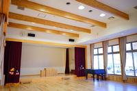 天井に木の梁が見える広々とした部屋に、カーテンで囲まれたステージと黒いピアノが設置されている写真