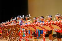 色とりどりの法被を着た子どもたちが舞台上に並び、紅白の装飾が施された太鼓を力強く叩いて一体感のある演奏を披露している写真