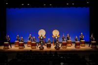 青い背景の舞台上で、多数の演奏者たちが大太鼓や中太鼓を使って一糸乱れぬ動きで力強く演奏を行っている和太鼓パフォーマンスの写真