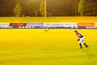 夜の野球場で、外野の芝生の上に立ち守備についている青いユニフォームの選手二人を、バックネット裏から撮影したスポーツの様子の写真