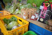 黄色と青のカゴの上にネギ、葉物野菜、米、豆などの農産物が整然と並べられている直売所の様子を写した写真