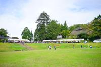 遠くに白いテントがいくつも並ぶ広場を背景に、広くて緑豊かな芝生広場で人々が散策している地域のイベント会場の写真