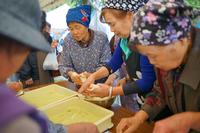 頭に手ぬぐいを巻いた年配の女性たちが屋根付きのテント内で手際よく餅を丸めている写真