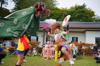 緑色の布で覆われた獅子舞と、カラフルな衣装と帽子をつけた子どもが対峙しながら踊っている、伝統芸能の披露場面の写真