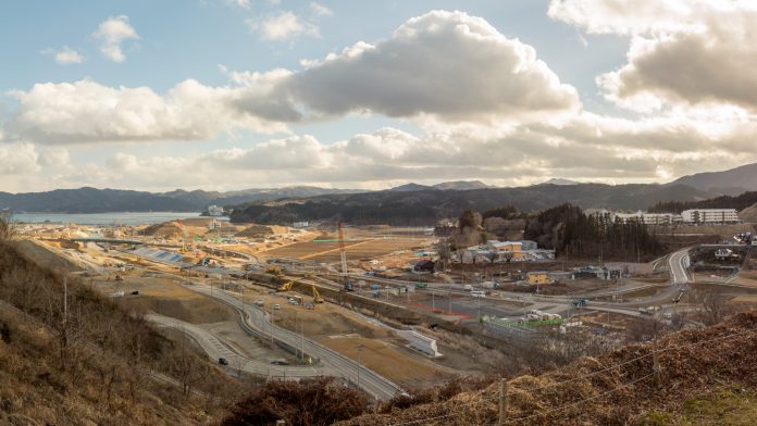 複数の道路や建物、建設機材が確認でき、背景には丘や山が広がり空は部分的に雲がかかっている建設現場を高台から写した写真