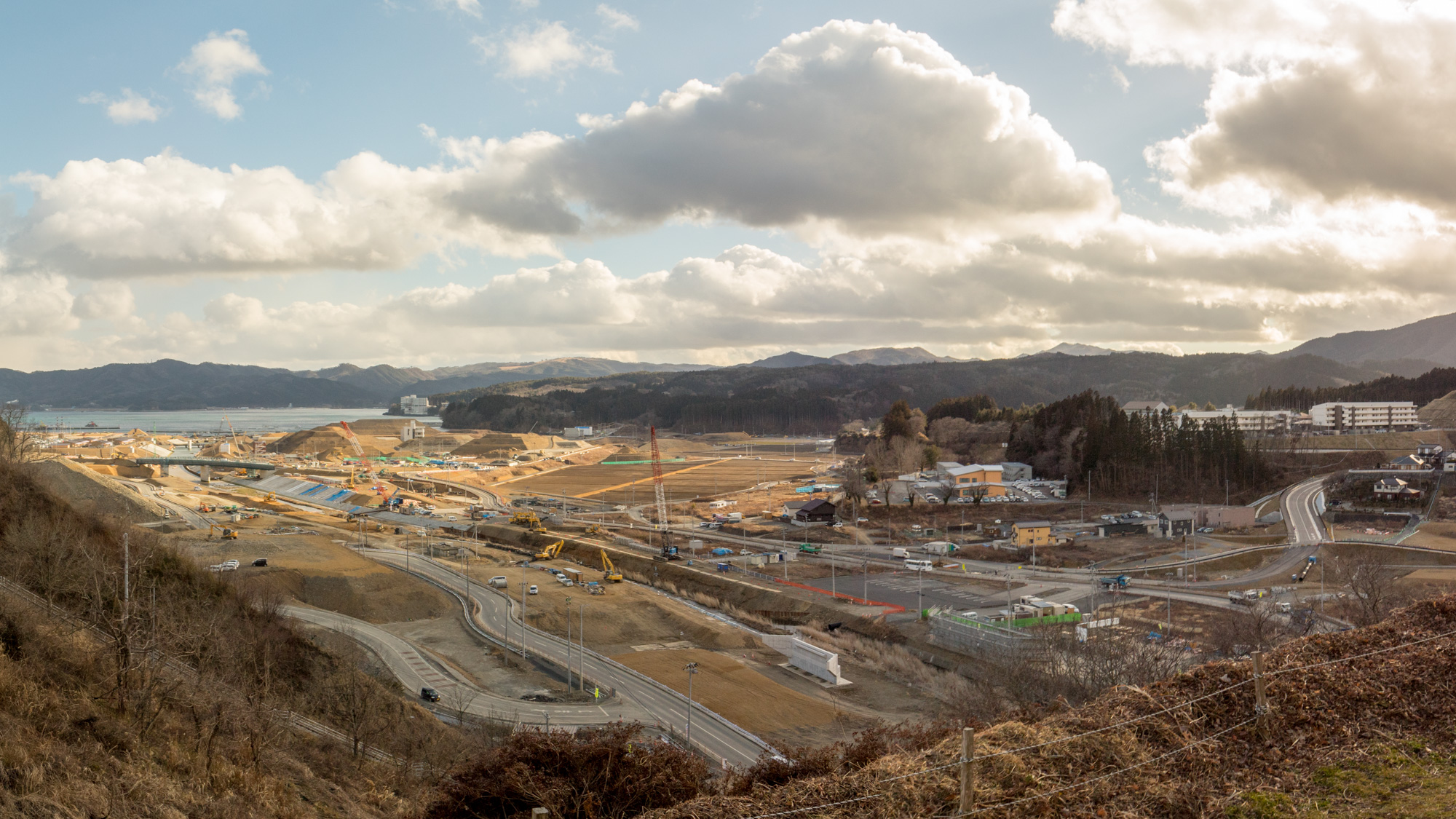 複数の道路や建物、建設機材が確認でき、背景には丘や山が広がり空は部分的に雲がかかっている建設現場を高台から写した写真