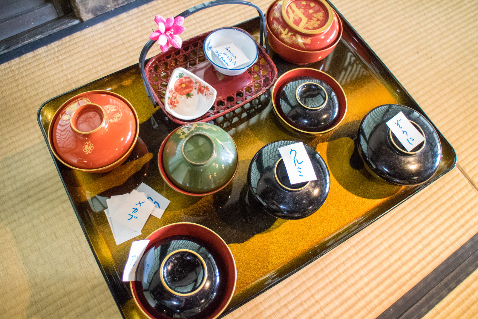 金色のお盆の上に蓋つきのお椀が7つ、2つの小皿が入った籠が1つ乗っており、料理名の紙が添えられている写真