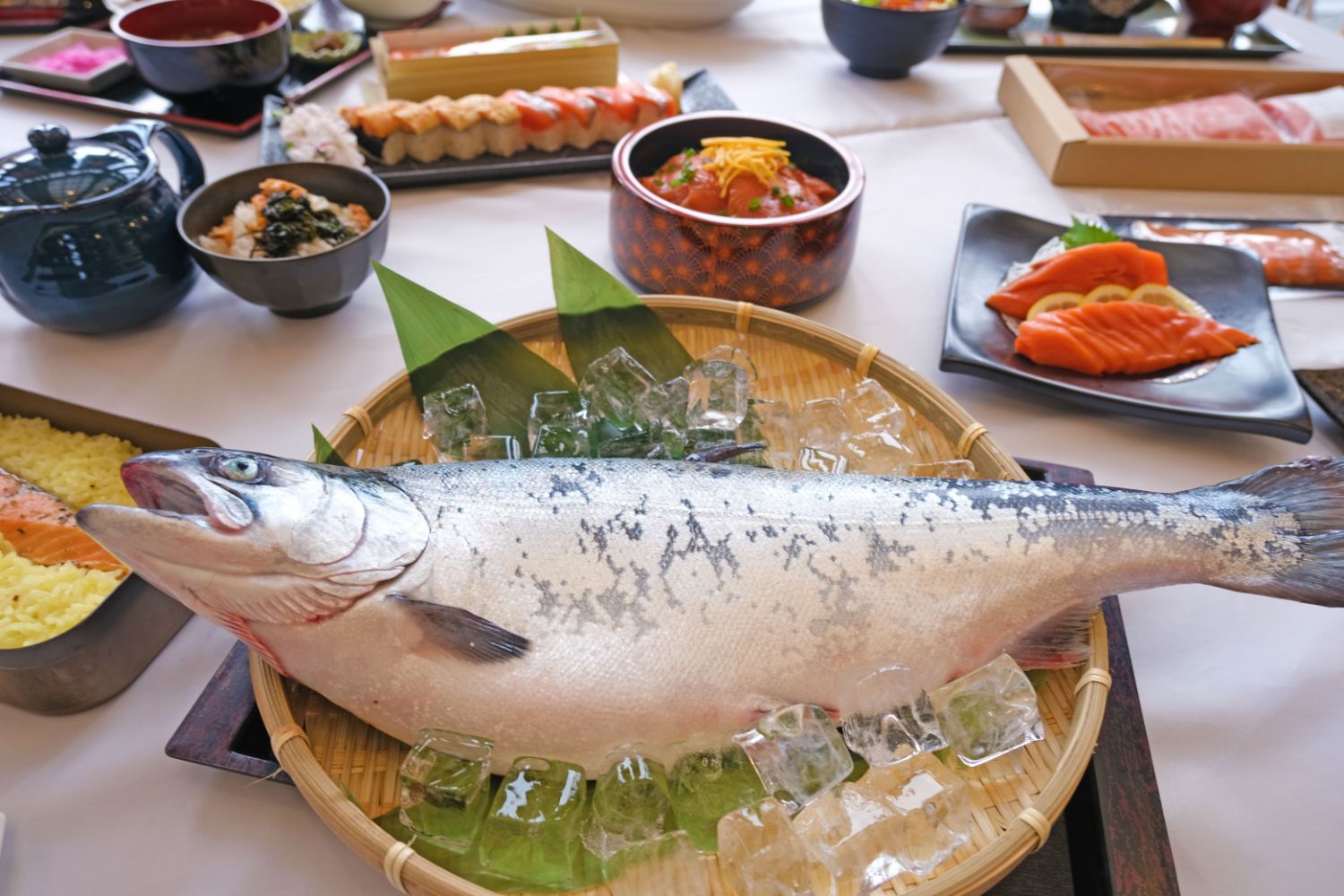 氷を敷き詰めた竹ざるの上に大きな鮭が丸ごと置かれ、その周りに刺身や寿司などの料理が並ぶテーブルのアップ写真