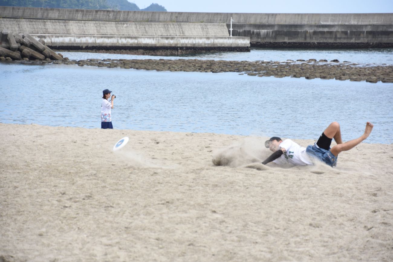 砂浜に倒れ込みながらディスクを追いかける選手と、それをカメラで撮影する女性の写真