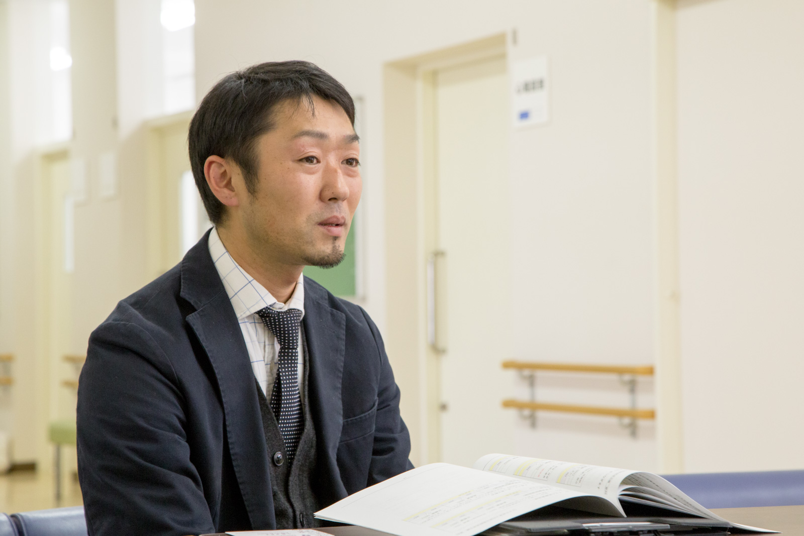 背景には白い壁と手すりのついた廊下、部屋のプレートが見える明るい室内で、開いた書類を前に座りながらインタビューをうけている男性の写真