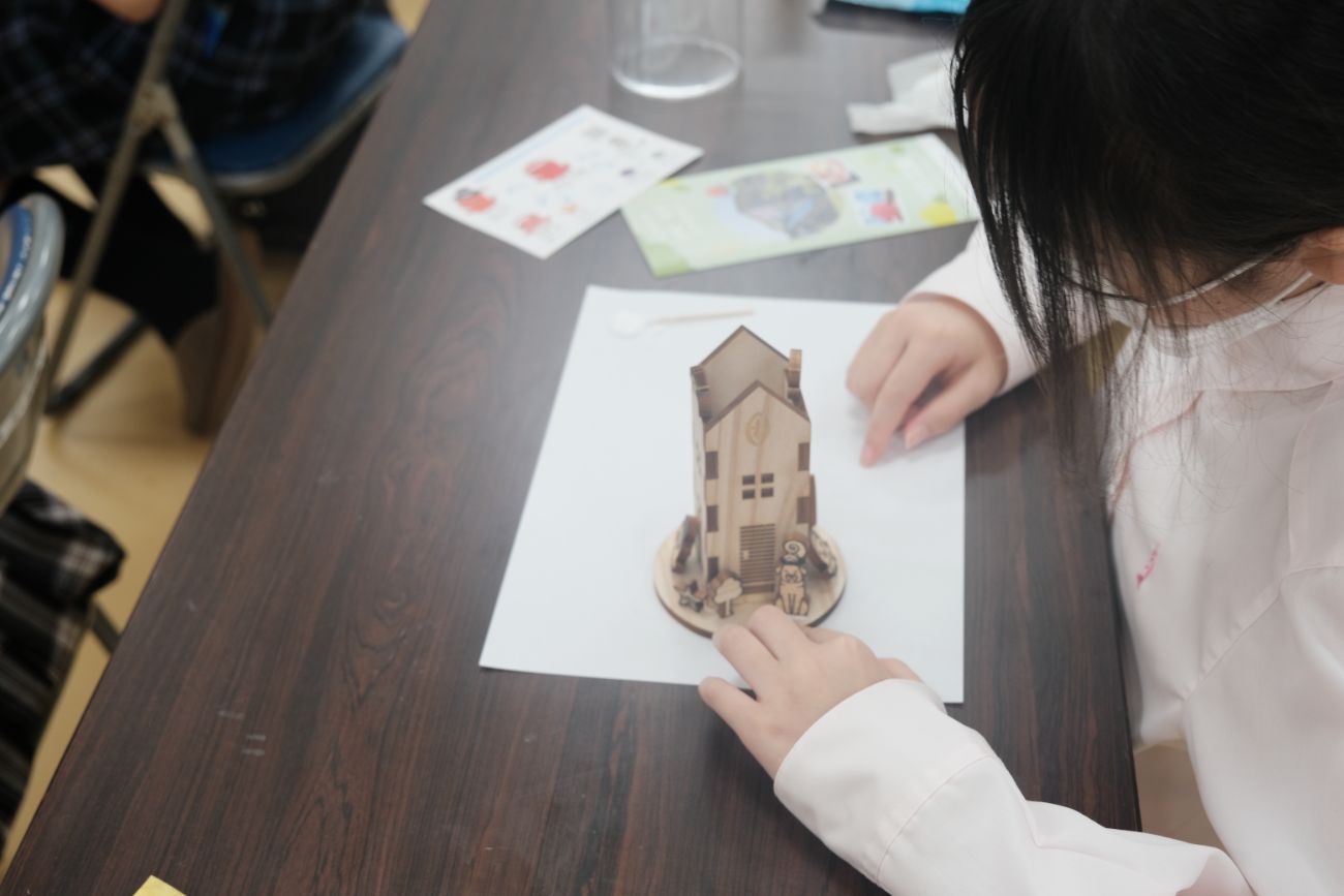 児童が紙の上に置かれた木製の小さな家の模型をじっと見つめて作業している様子の写真