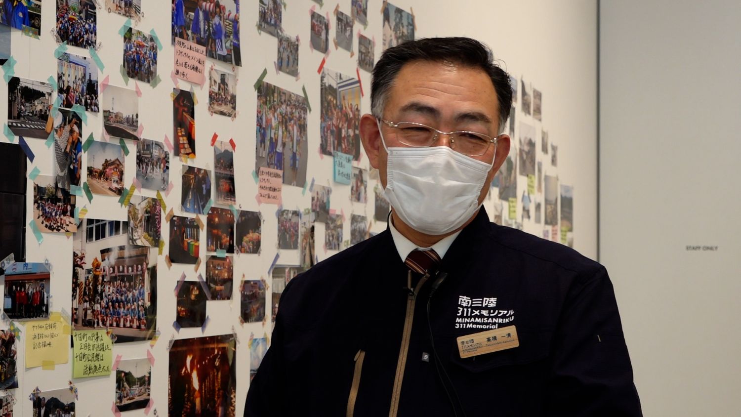 「南三陸311メモリアル」の制服を着た高橋一清さんがマスクを着用しながら、展示された写真の前で写っている写真