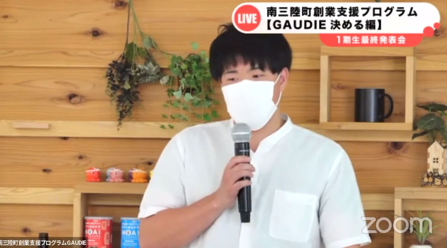 白いシャツを着た男性がマスクを着けてマイクを持ち、木目の背景の前で南三陸町創業支援プログラムGAUDIEの発表会で話している様子の写真