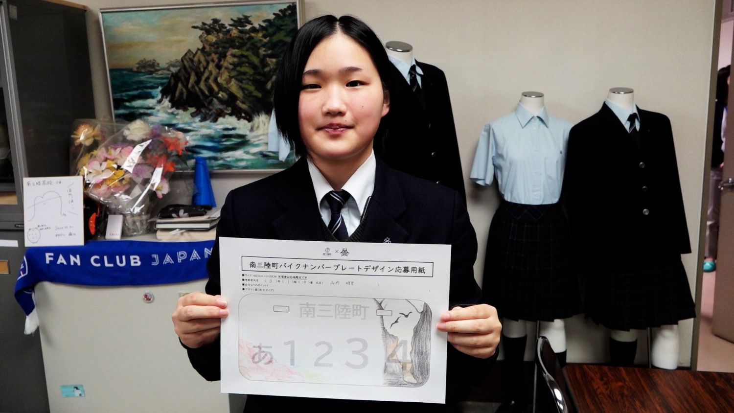 自作のナンバープレート原案をカメラに見せる女子高校生の写真