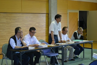 会議中に前方の机に座っていた千葉正海さんが立ち上がって開会のあいさつをしている様子の写真