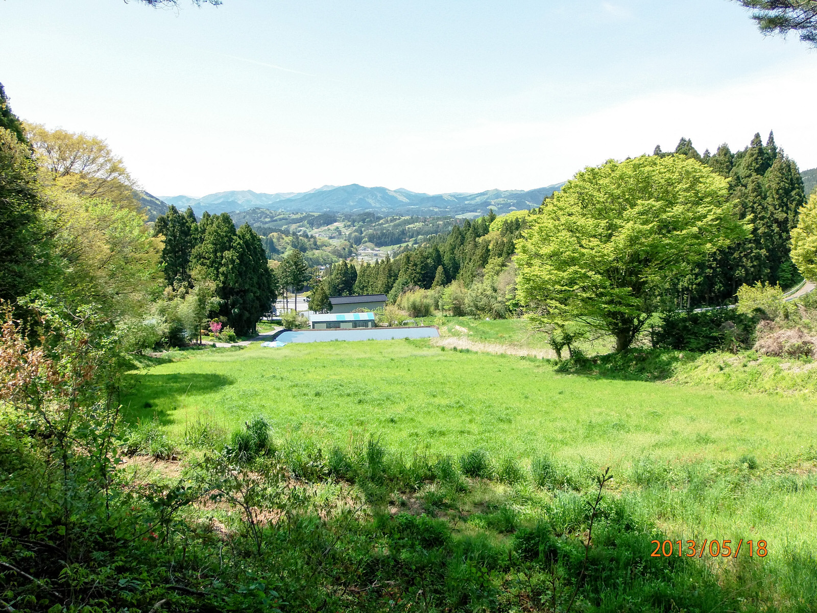 新緑に包まれた山々と広々とした草原の奥に小さな建物が見える、自然豊かな里山風景の写真