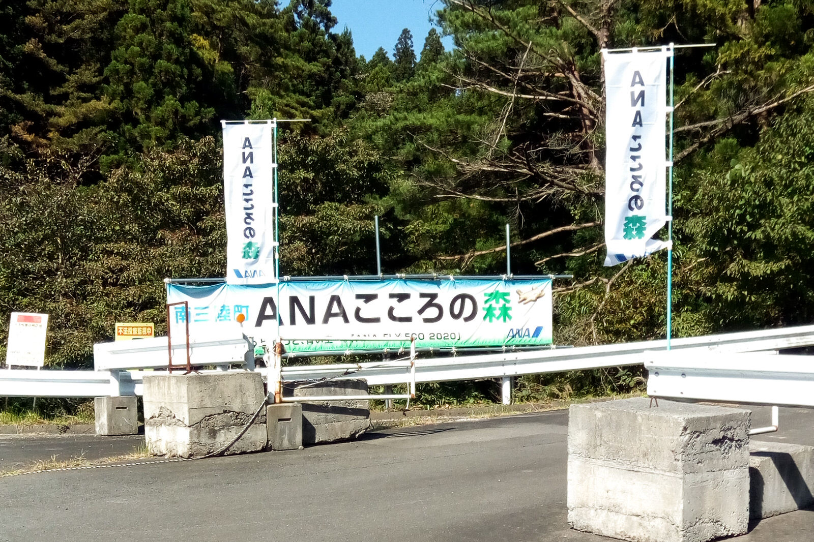 ガードレールが設置された山林の入り口に「ANAこころの森」と書かれた横断幕と旗が掲げられている写真