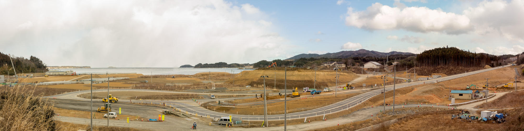 現場には複数の建設車両や機材が点在しており、ショベルカーやトラックなどが確認でき、背景には丘や山が広がり、右側にはいくつかの建物も見える工事中の沿岸風景のパノラマ写真