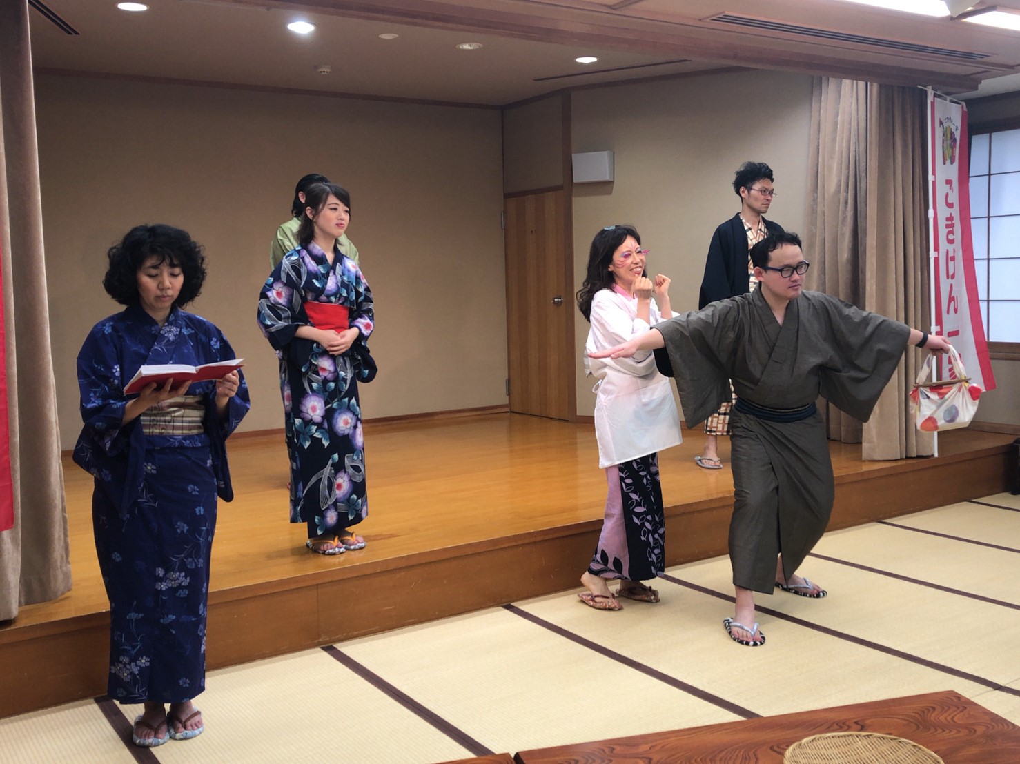舞台上とその前で6人の男女が浴衣や割烹着姿で演技をしている様子の写真