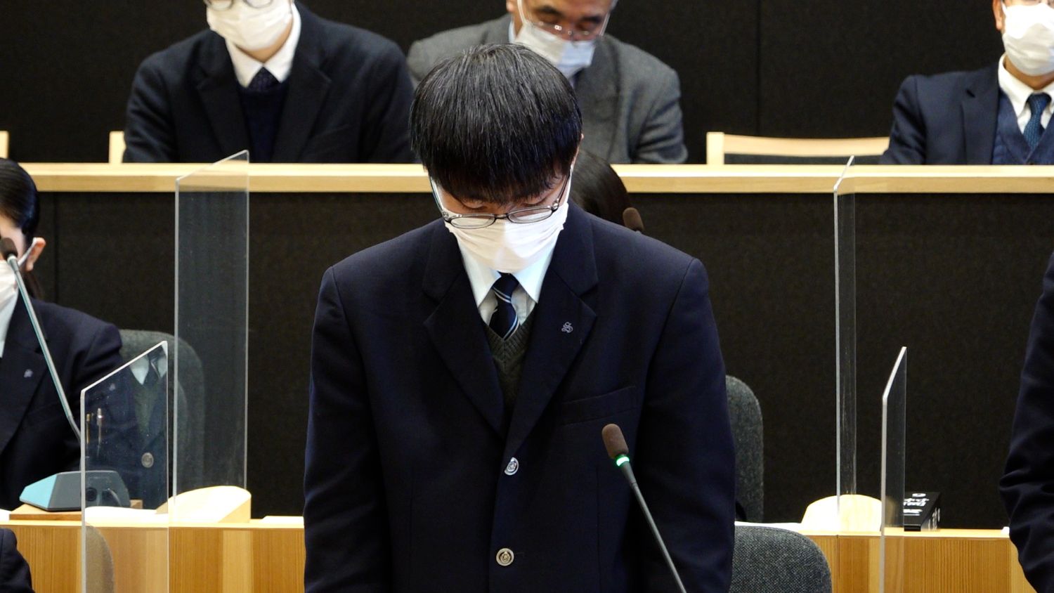 透明なアクリル板が設置された議場で、制服姿の男子生徒が深く頭を下げて発言の場面に臨んでおり、背後にはスーツ姿の大人たちが静かに見守っている写真