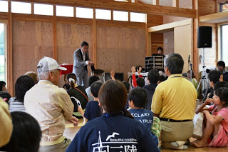室内のステージ前に子どもたちが集まり、スーツ姿の男性が語りかけるように手を挙げている子どもたちに応えている催し中の写真