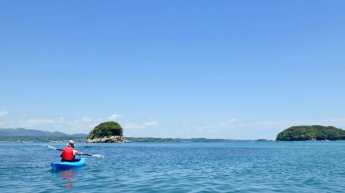 山と青空が見える海の上で、青いカヤックに乗った赤いライフジャケットの人物が穏やかな海の上を一人で漕ぎ、緑に覆われた小さな島に向かって進んでいる写真
