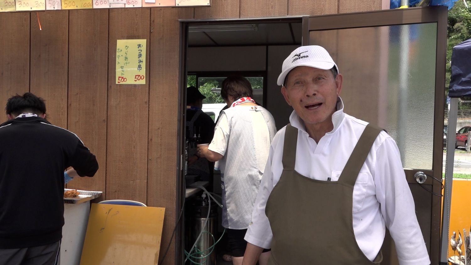 背後では数人が忙しそうに調理や作業をしている木目調の壁に囲まれた店の前で、白い帽子とシャツに茶色のエプロンを着けた年配の男性が笑顔で立っている写真