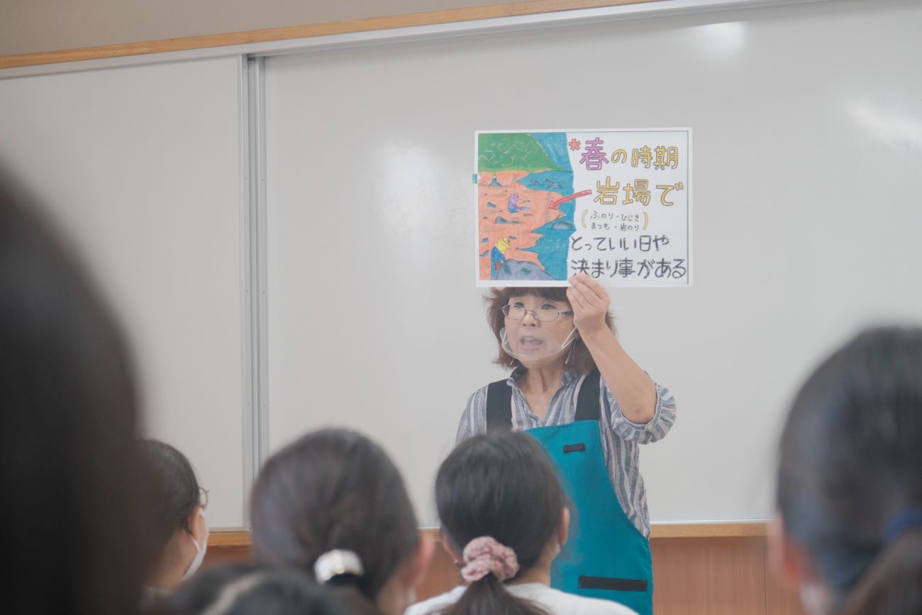 女性が「春の時期 岩場で」というタイトルの紙を掲げ、子どもたちに説明をしている写真