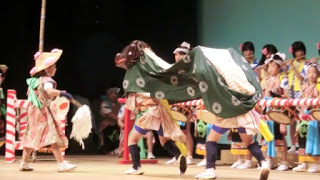 色鮮やかな伝統衣装を身にまとった演者たちが、白い房飾りを持つ人物や緑色で白い模様の衣装をまとった獅子役の踊り手を中心に、太鼓や笛の演奏とともに舞台上でにぎやかに披露している入谷打囃子（いりやうちばやし）の郷土芸能発表の写真